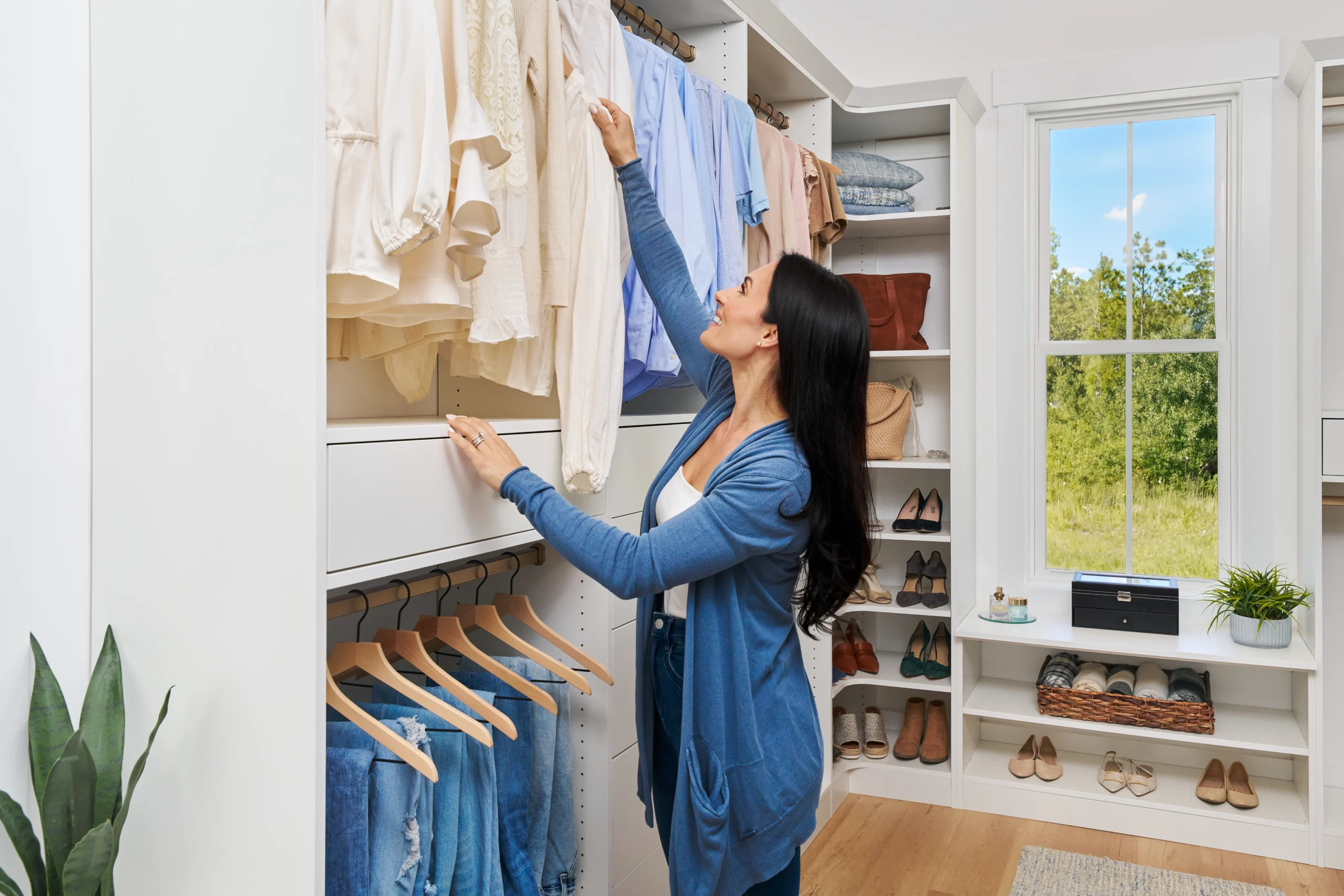 A woman in a white closet with organized clothes on shelves and hanging rods, reaching for a shirt