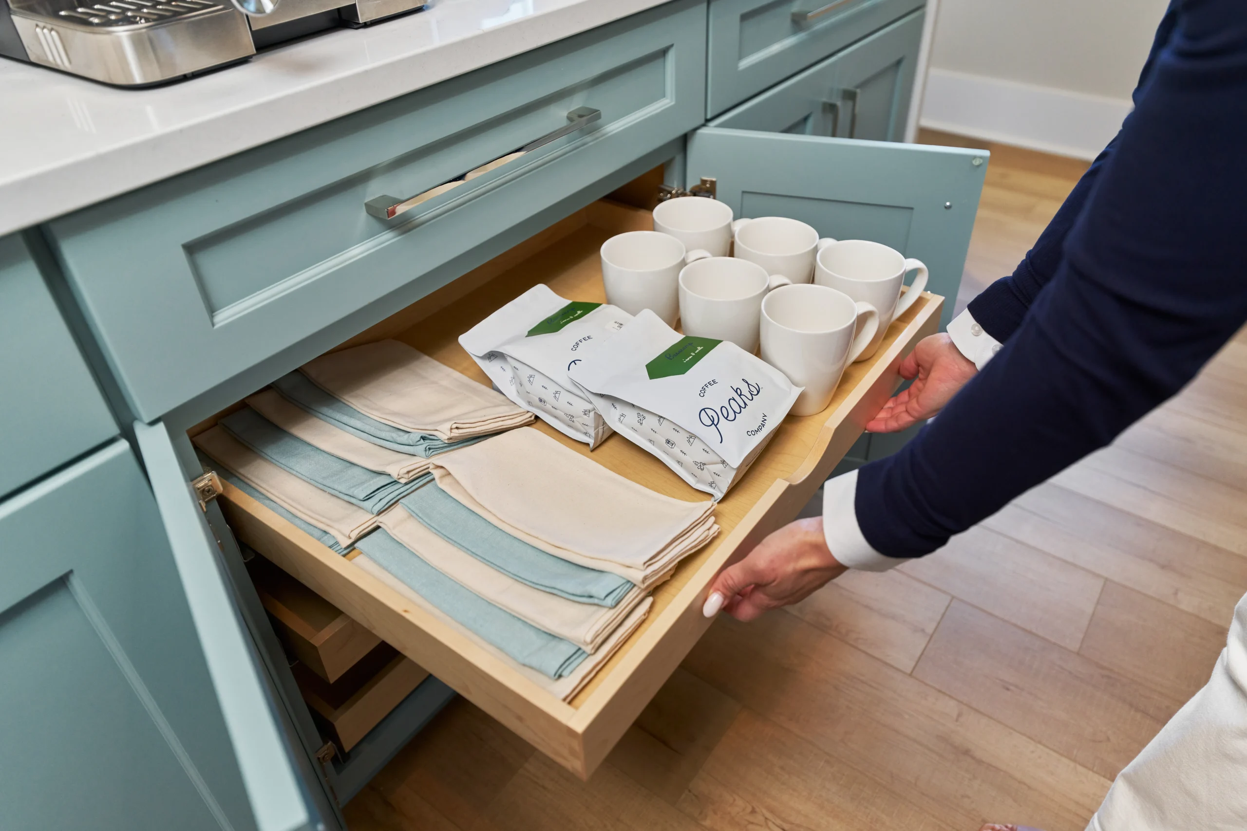 A hand pulls out a drawer filled with white mugs, coffee bags, and neatly folded cloth napkins in a modern kitchen